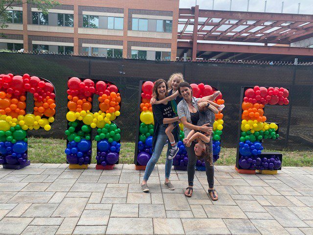 Nicole G. with wife, daugther and son standing in front of Pride sign at Vanguard campus