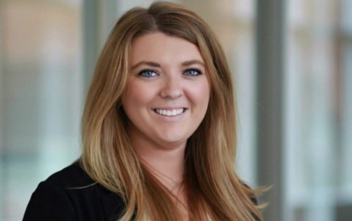 Rachel C. is smiling confidently in her professional headshot, wearing a black sweater over a white top. The background is blurred, showing what appears to be an office setting with large windows and natural light.