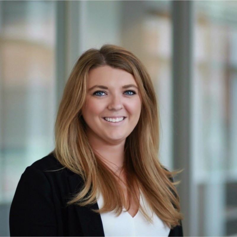 Rachel C. is smiling confidently in her professional headshot, wearing a black sweater over a white top. The background is blurred, showing what appears to be an office setting with large windows and natural light.